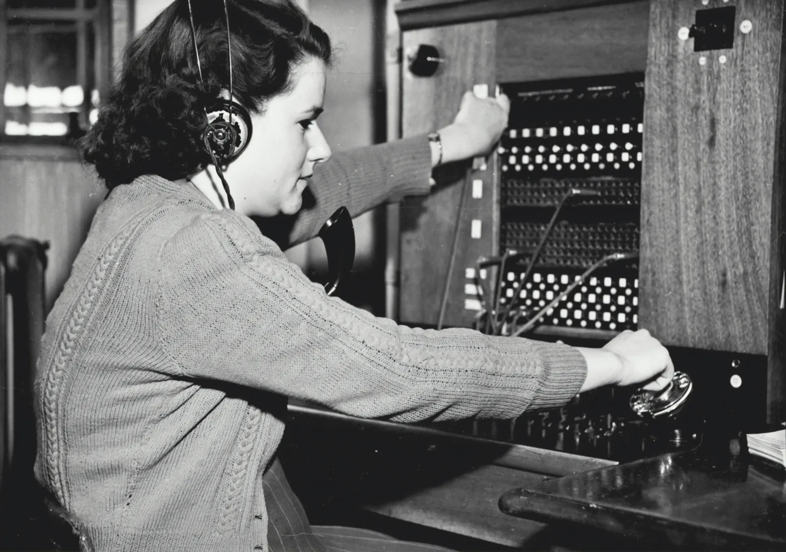 A 1940s switchboard operator routing tangled cables on a wooden patch panel: too many wires to plug, too few hands to plug them.