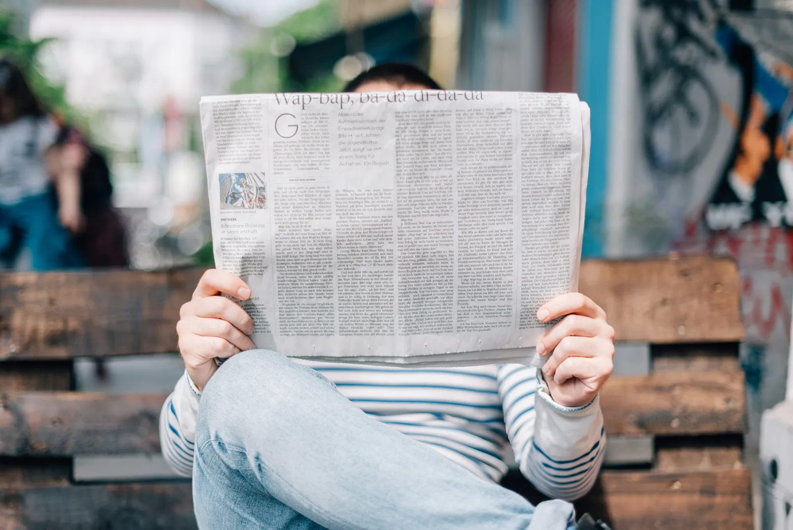 A person on a bench reads a multi-column newspaper that hides their face, standing for a reader who cannot see how the column layout is being scrambled.